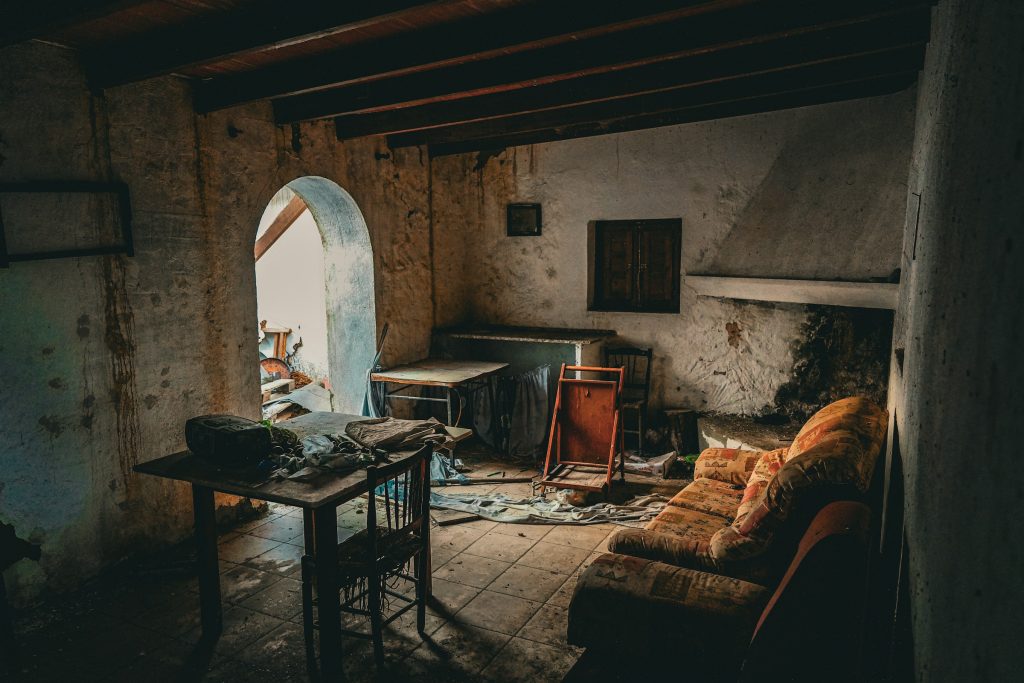 Before: a run-down cottage room with dark beams, an arched doorway, a hooded fireplace, stained walls and old furniture