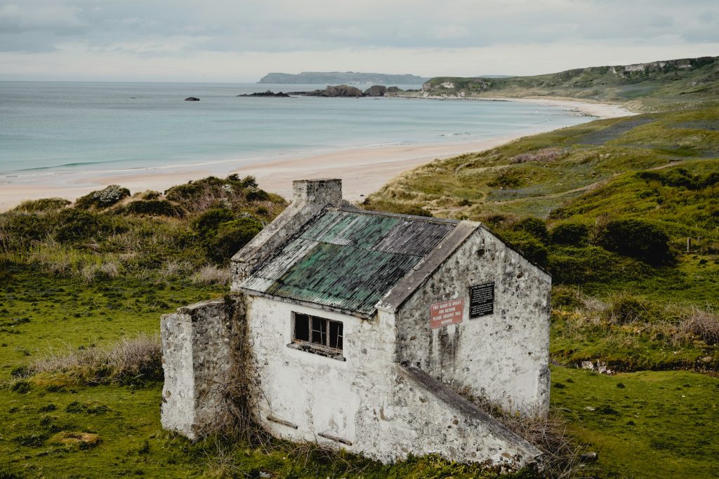 Before: a derelict whitewashed cottage with a mossy, broken roof on a grassy hillside above a sandy beach