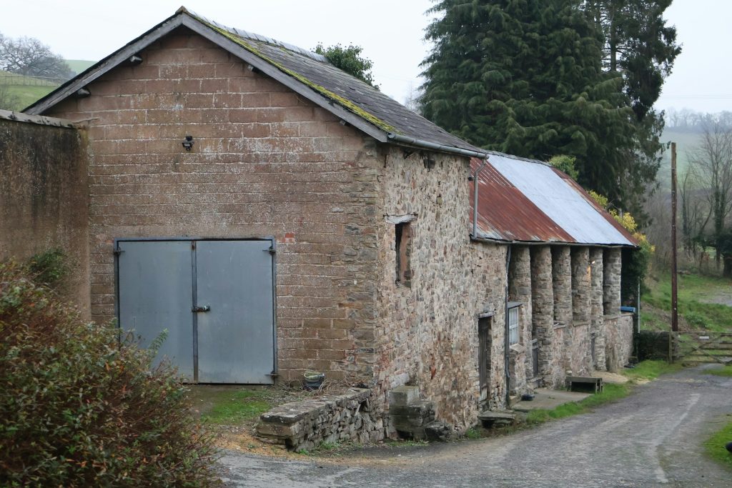 Before: a stone and block farm outbuilding with a mossy slate roof, a rusty corrugated lean-to and grey metal doors